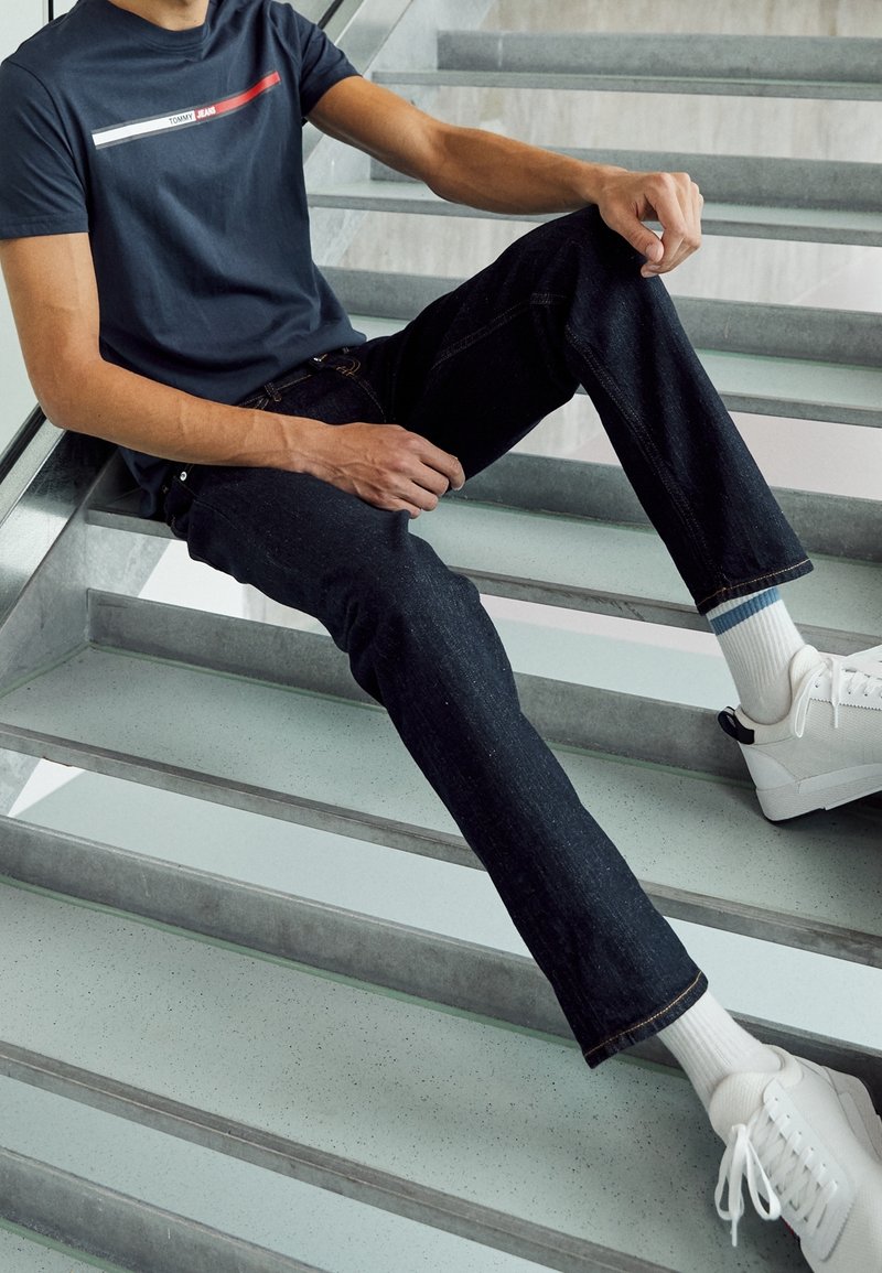 Námořnické bavlněné tričko s horizontálním červeným a bílým vzorem, spárované s tmavými džínami a bílými teniskami se šedými podrážkami.