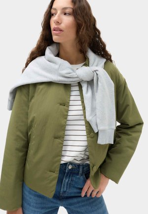 Ung kvinde med langt krøllet hår iført grøn jakke over en stribet skjorte og blå jeans, med en grå sweatshirt bundet omkring skuldrene.