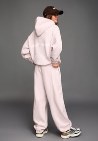Svetloružová nadmerná mikina s veľkou kapucňou a predným vreckom, skombinovaná s rovnako sfarbenými zužujúcimi sa teplákmi. Hnedá čiapka a farebné tenisky.