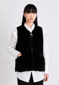 Schwarzer Kunstpelz-Weste mit hohem Kragen, durchgehendem Reißverschluss und seitlichen Taschen, getragen über einem weißen Hemd mit Knopfleiste. Die Textur ist weich und plüschig.