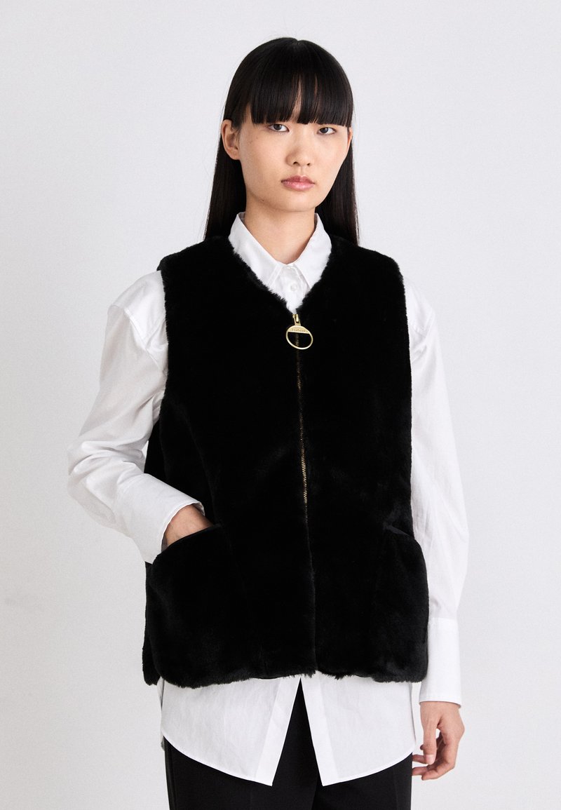 Schwarzer Kunstpelz-Weste mit hohem Kragen, durchgehendem Reißverschluss und seitlichen Taschen, getragen über einem weißen Hemd mit Knopfleiste. Die Textur ist weich und plüschig.