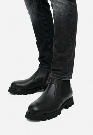 Schwarze Leder-Stiefeletten mit dicken Sohlen, getragen zu dunklen, ausgewaschenen Jeans vor weißem Hintergrund.