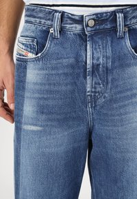 Lichtblauwe denim jeans met een relaxte pasvorm, voorzakken en een klein label zichtbaar in de linkerzak. Gestructureerde stof met vervagingdetails.