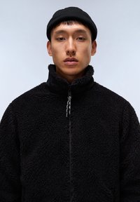 Giacca in fleece nera con collo alto, chiusura con zip e tessuto testurizzato. Indossata con un berretto nero, su uno sfondo grigio chiaro.