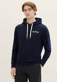 Felpa navy in cotone misto con tasca frontale, colletto con lacci, logo bianco e polsini a coste. Il modello si trova davanti a uno sfondo neutro.