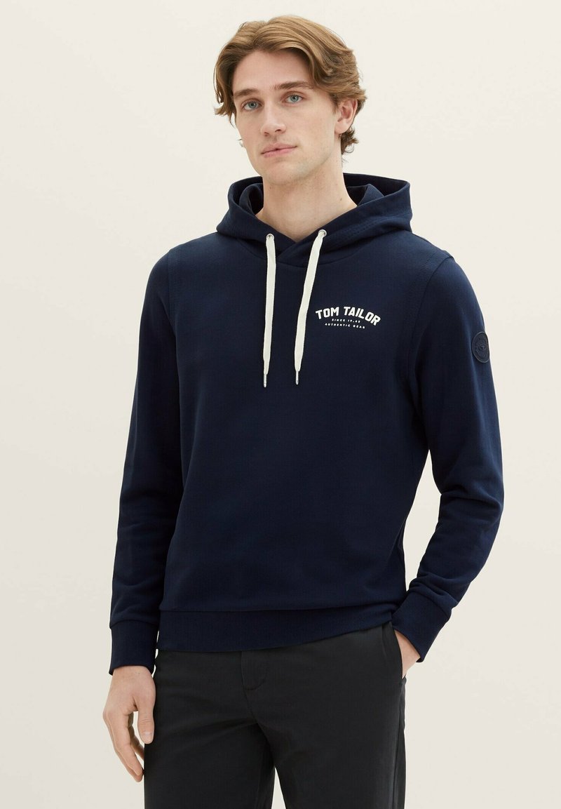 Felpa navy in cotone misto con tasca frontale, colletto con lacci, logo bianco e polsini a coste. Il modello si trova davanti a uno sfondo neutro.