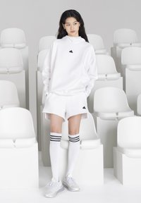 Biela mikina s kapucňou a čiernym logom, zodpovedajúce biele šortky a biele ponožky po kolená s čiernymi pruhmi. Strieborné športové topánky dopĺňajú tento outfit.