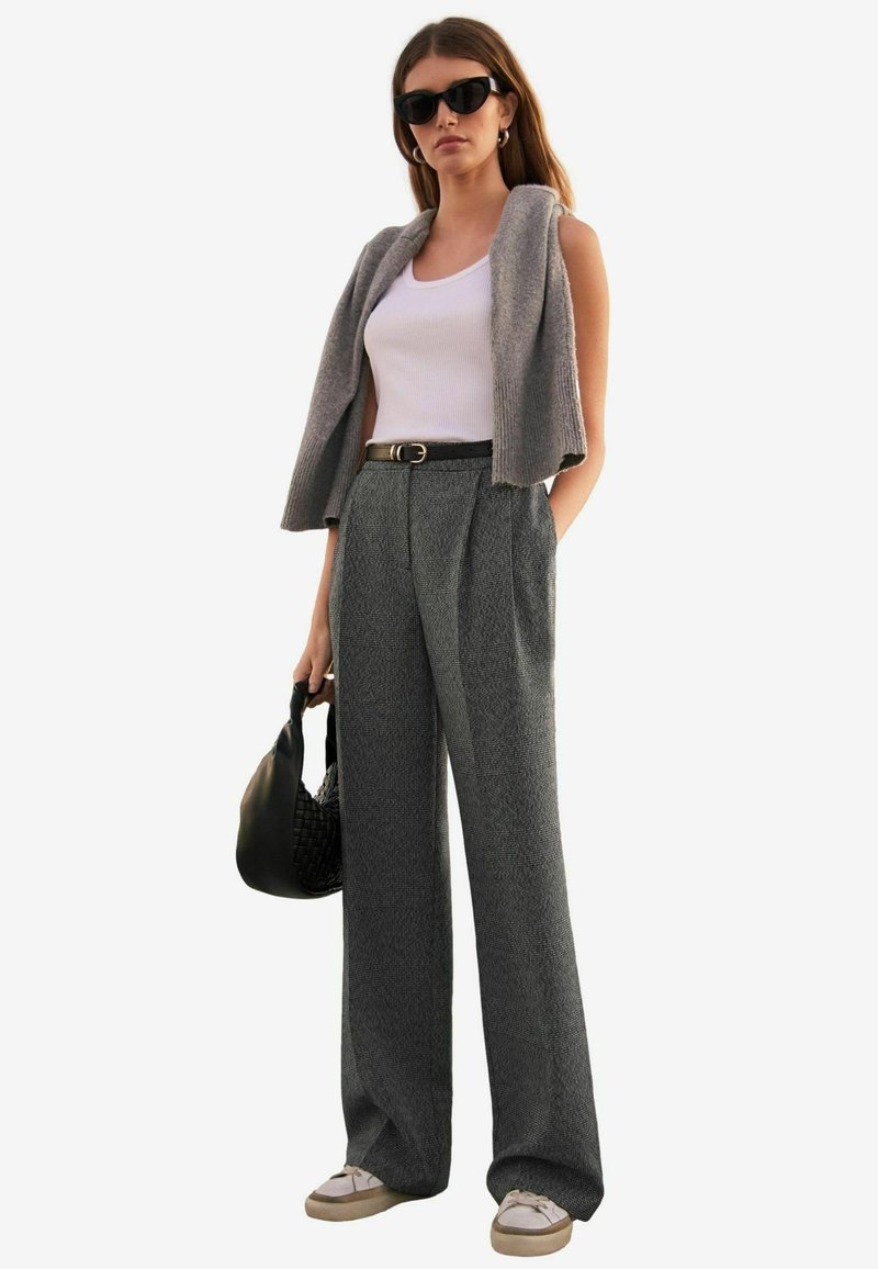Gilet grigio di maglia sopra una canotta bianca, pantaloni a quadri neri e grigi a vita alta, borsa nera e sneakers beige con lacci bianchi.