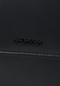 Schwarze Lederoberfläche mit strukturiertem Finish, das das geprägte "Calvin Klein"-Logo und kontrastierende Nähte entlang der Kante zeigt.