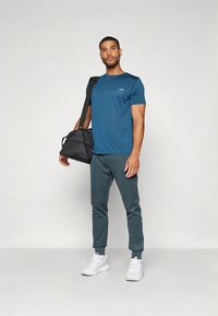 Tee-shirt sportowy w kolorze niebieskozielonym z krótkimi rękawami, zestawiony z ciemnozielonymi joggerami z bocznymi paskami. Czarna torba podróżna i białe sneakersy.