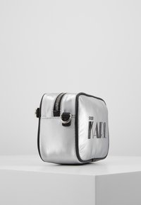 Silberne Satin-Schminktasche mit schwarzen Akzenten, rechteckige Form, Reißverschluss, mit dem Logo "STUDIO KARL" in fetten schwarzen Buchstaben.