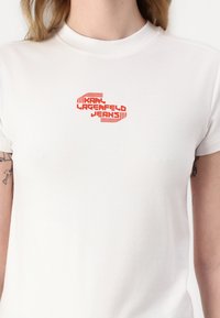 Bílá bavlněná tričko s výrazným červeným grafickým logem "KARL LAGERFELD JEANS" uprostřed, s krátkými rukávy a kulatým výstřihem.