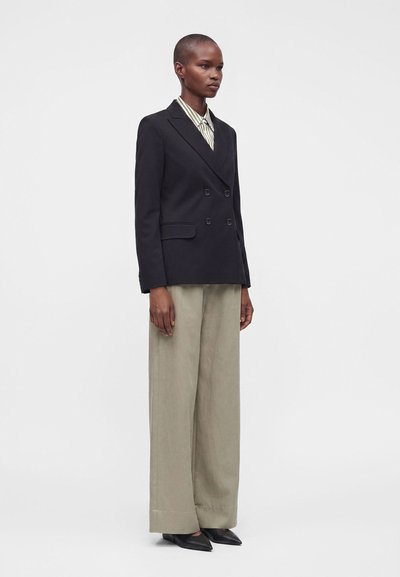 Mannequin portant un blazer noir croisé, chemise rayée, pantalon beige large et chaussures noires pointues, debout devant un fond uni.