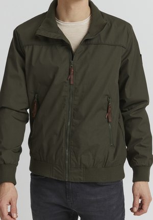 Mand iført olivengrøn lynlåsjakke med ribbet krave, manchetter og kant, over beige skjorte og mørke jeans, stående mod ensfarvet baggrund.