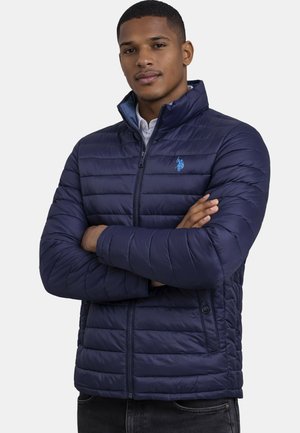 Man with crossed arms wearing a navy blue quilted jacket with a high collar and a small blue horse logo on the chest.