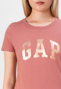 Young person wearing dusty pink T-shirt with large metallic gold letters "GAP" across the chest, visible from shoulders to mid-torso.