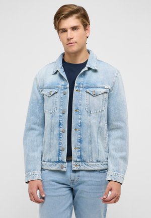 Młody mężczyzna ubrany w jasnoniebieską dżinsową kurtkę i pasujące jeansy, na ciemnogranatowej koszulce, stojący na tle jasnego, jednolitego tła.