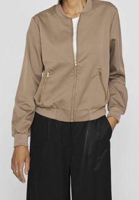 Femme portant une veste beige à fermeture éclair dorée et un pantalon noir taille haute, avec une main dans la poche et des bagues aux doigts.