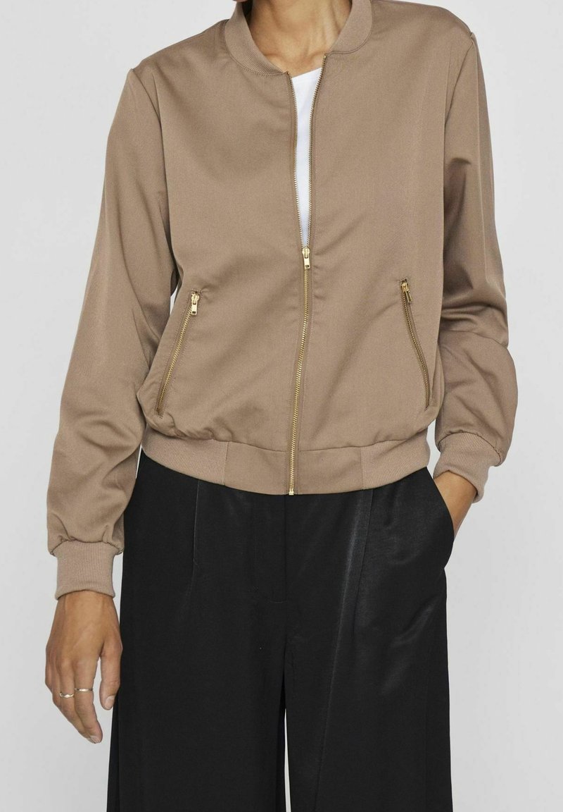 Femme portant une veste beige à fermeture éclair dorée et un pantalon noir taille haute, avec une main dans la poche et des bagues aux doigts.