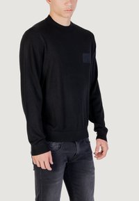 Maglione nero a maniche lunghe con collo rotondo, dotato di una tasca sul petto texturizzata e polsini a coste, abbinato a jeans scuri. Tessuto morbido e liscio.