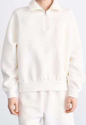 Persona que lleva un suéter blanco de manga larga y cuello con cremallera de un cuarto, combinado con pantalones blancos a juego, frente a un fondo liso.