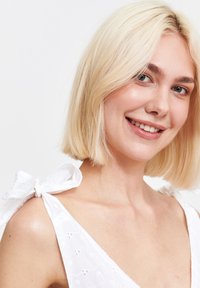 Uśmiechnięta kobieta z blond bobem ma na sobie białą sukienkę z kokardkami na ramionach; stoi nieco zwrócona w lewo na jasnym tle.