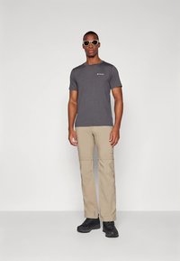 Pilka trumpomis rankovėmis marškinėliai su logotipu, derinami su smėlio spalvos nuimamo ilgio krovinių kelnėmis ir juodais tvirtais batais, pozicijoje prieš paprastą foną.