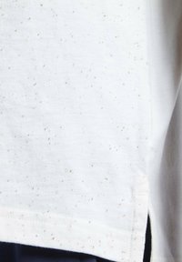 Tissu blanc avec de petites taches grises, couture visible et fente latérale sur des vêtements sombres en dessous.