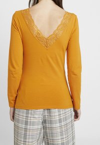 Long-sleeve, mustard-orange top with a deep V-back featuring intricate lace trim. Smooth fabric, fitted silhouette, paired with plaid trousers.
