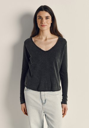 Giovane donna con capelli lunghi e scuri che indossa una maglietta a maniche lunghe grigio scuro con scollo a V e jeans azzurri, in piedi contro un muro bianco sporco uniforme.