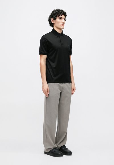 Melnā īsrokas polo krekls ar apkakli, kopā ar gaiši pelēkām biksēm un melniem sporta apaviem, kas atspoguļo vienkāršu, mūsdienīgu dizainu.