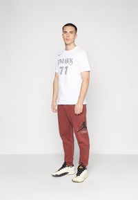 Bjela košulja kratkih rukava s sivim grafičkim natpisom "MAVS 77" i crvene trenirke s crnim Jordan logom. Crno-bijele sportske tenisice.