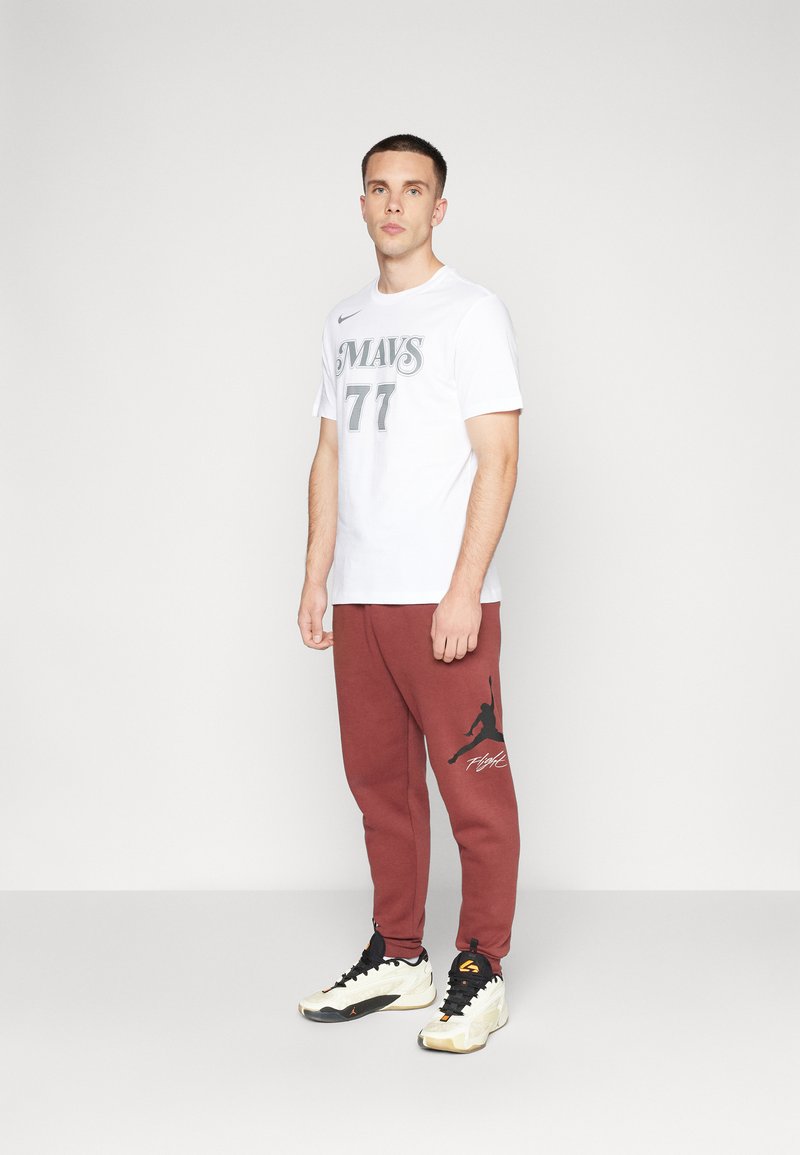 Weißes Kurzarmhemd mit grauem Grafikdesign "MAVS 77" und roten Jogginghosen mit schwarzem Jordan-Logo. Schwarz-weiße Sportschuhe.