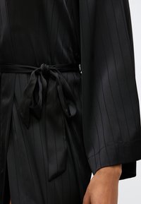 Μαύρη σατέν ρόμπα με κατακόρυφες ραβδώσεις, φαρδιά μανίκια και ζωνάκι που δένει στη μέση. Λευκή υφή με διακριτική λάμψη και χαλαρή σιλουέτα.