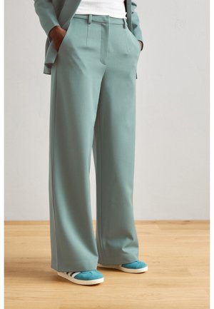 Person wearing loose-fitting light teal trousers, white top, matching teal sneakers with white stripes, standing on wooden floor.