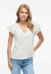 Blusa blanca de cuello en V con mangas cortas y acampanadas que presenta detalles bordados en verde. Confeccionada en una tela ligera con un acabado texturizado.