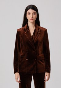 Blazer double boutonné en velours marron avec revers crantés, deux poches avant et deux boutons. Texture lisse et coupe ajustée.