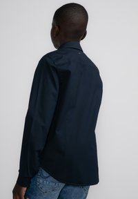 Marineblauwe langearmige shirt met een kraag, gemaakt van een gladde stof, met een rugpand en een subtiele merkdetail nabij de nek.
