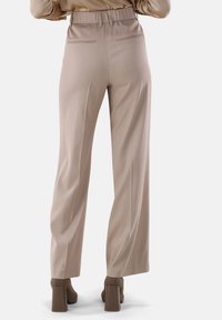 Weite Hosen aus hellbeigem Stoff mit strukturiertem Oberflächen, ausgestattet mit einem hohen elastischen Bund und zwei Gesäßtaschen. Braune Schuhe mit Blockabsatz.