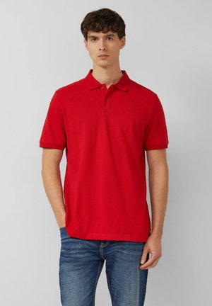 Mann trägt ein schlichtes, knallrotes, kurzärmeliges Polohemd und blaue Jeans, steht vor einem schlichten hellgrauen Hintergrund.