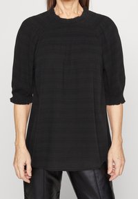 Blouse noire avec une finition texturée, dotée d'un col rond et de manches bouffantes trois-quarts avec des poignets froncés. Design à coupe droite.
