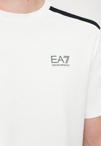 White cotton t-shirt featuring a crew neck, black shoulder accents, and a logo print on the left chest in gray. Smooth texture.