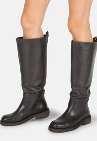 Botas altas de cuero negro hasta la rodilla con punta redondeada y tacón apilado de madera, que presentan un acabado suave y texturizado, además de una tira en la parte superior para facilitar su uso.