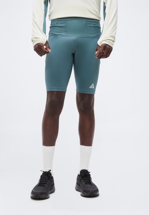 Homme portant un short de sport bleu sarcelle, une chemise blanche à manches longues, des chaussettes blanches et des baskets noires, debout devant un fond uni.
