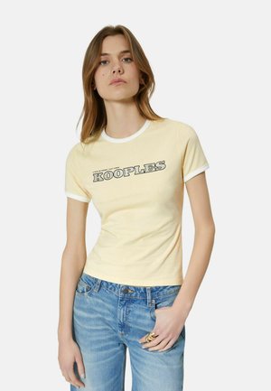 Vrouw draagt een lichtgele aansluitende T-shirt met witte bies en de tekst "KOOPLES", gecombineerd met lichtblauwe spijkerbroek en gouden ringen aan de rechterhand.