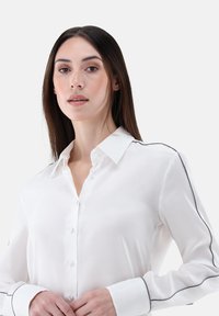 White button-up shirt with a pointed collar, featuring contrasting dark piping along the shoulders and cuffs. Smooth, lightweight fabric.