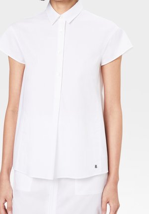 Vrouw draagt een wit kort mouw blouse met een klein zwart logo vlak bij de zoom, gecombineerd met een witte rok of broek.