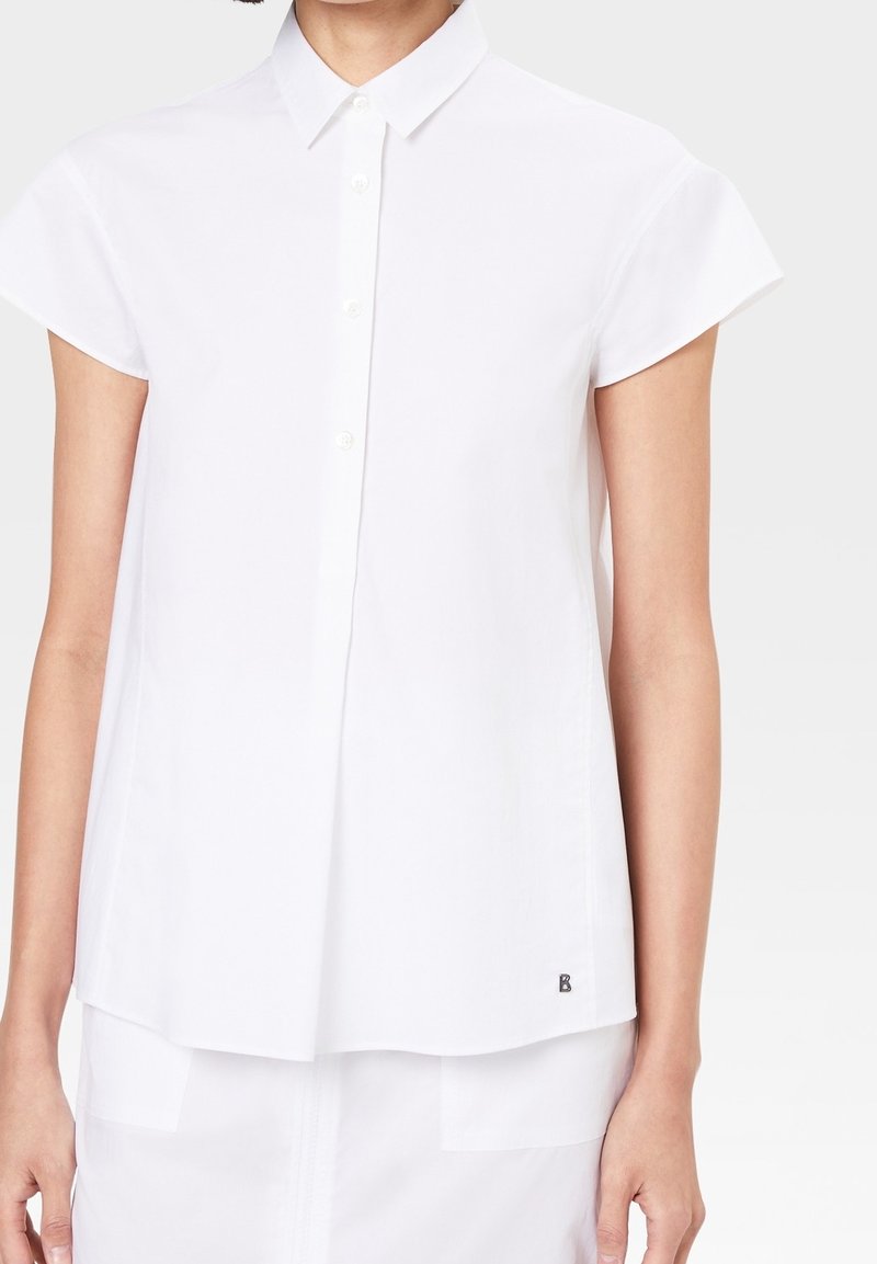 Femme portant une chemise blanche à manches courtes avec un petit logo noir près de l'ourlet, associée à une jupe ou un pantalon blanc.