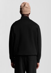 Svart sweatshirt med avslappnad passform, ribbad krage och långa ärmar. Bärs tillsammans med en ljusbrun stickad mössa. Slät textur och minimalistisk design.