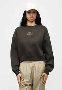 Γυναίκα που φοράει σκούρο γκρι φούτερ New Balance, μπεζ παντελόνι με ελαστική μέση και άσπρο καπέλο του μπέιζμπολ LA, στέκεται μπροστά σε ένα απλ ό φόντο.